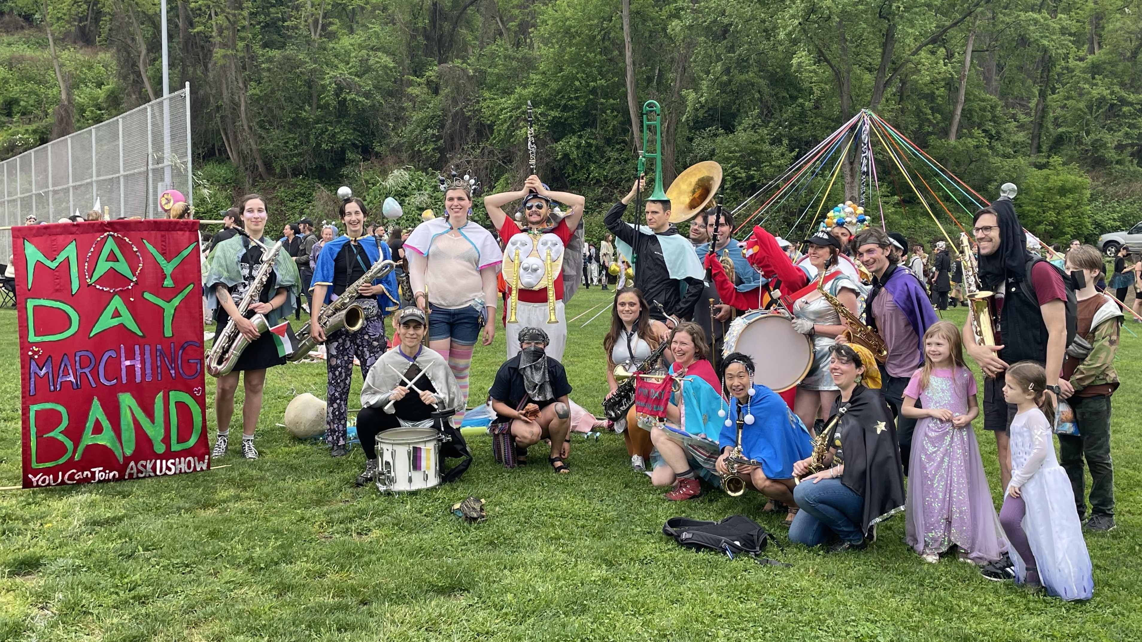 May Day Marching Band poses for a picture in front
                  of a maypole and other festive elements. A banner
                  in the left side of the frame reads "You can
                  join ASK US HOW"