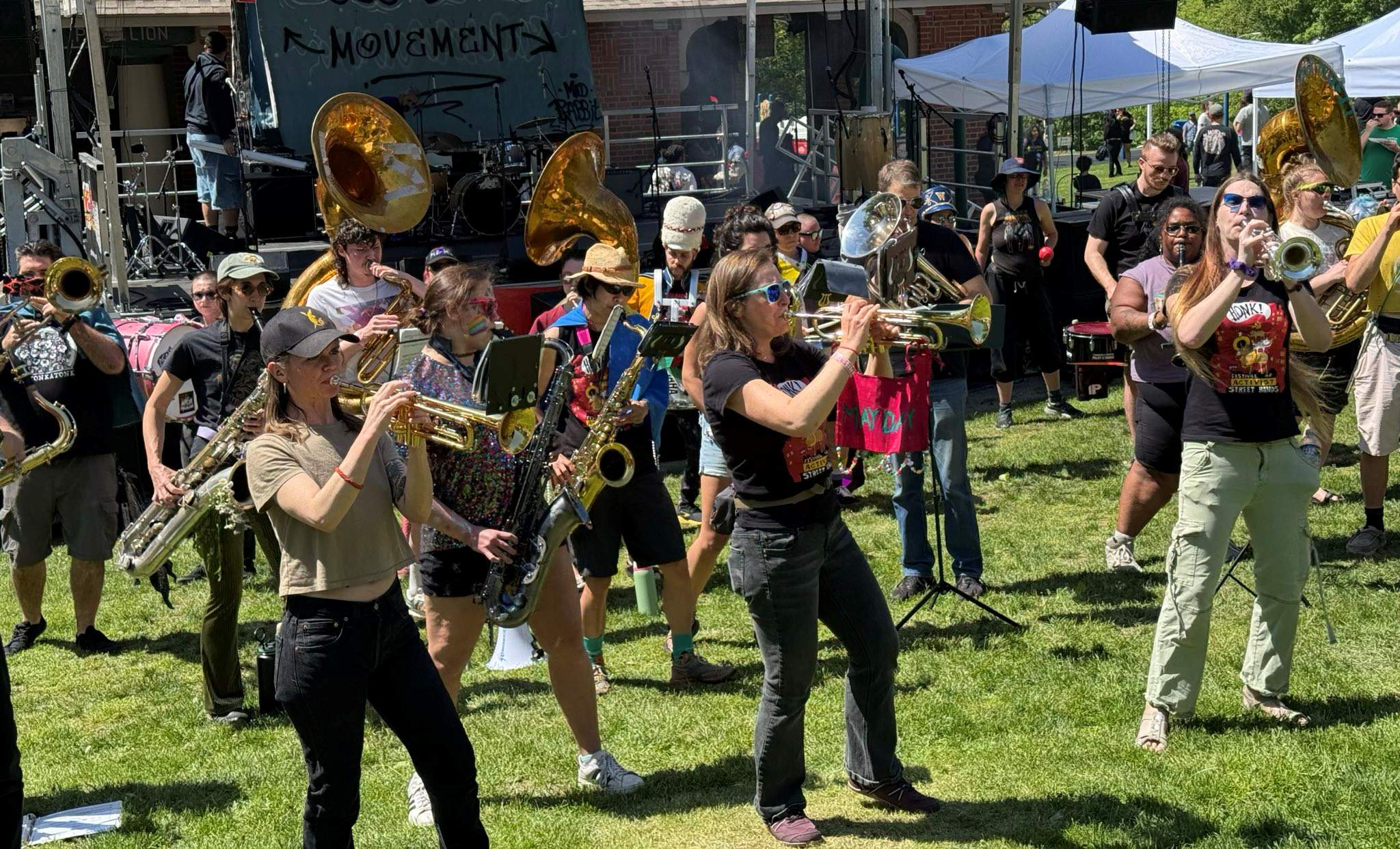 May Day Marching Band performs in a field in front
                of a stage with a graffiti-style backdrop saying
                "MOVEMENT"